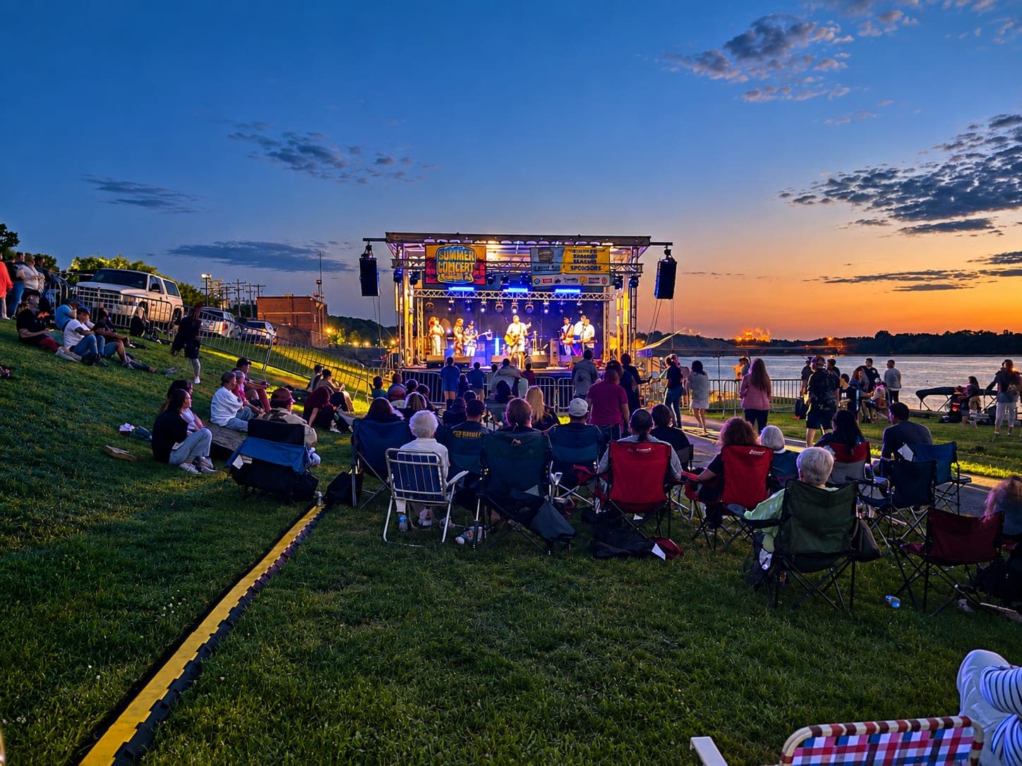 Outdoor stage with a band playing at dusk, audience on the lawn by the river